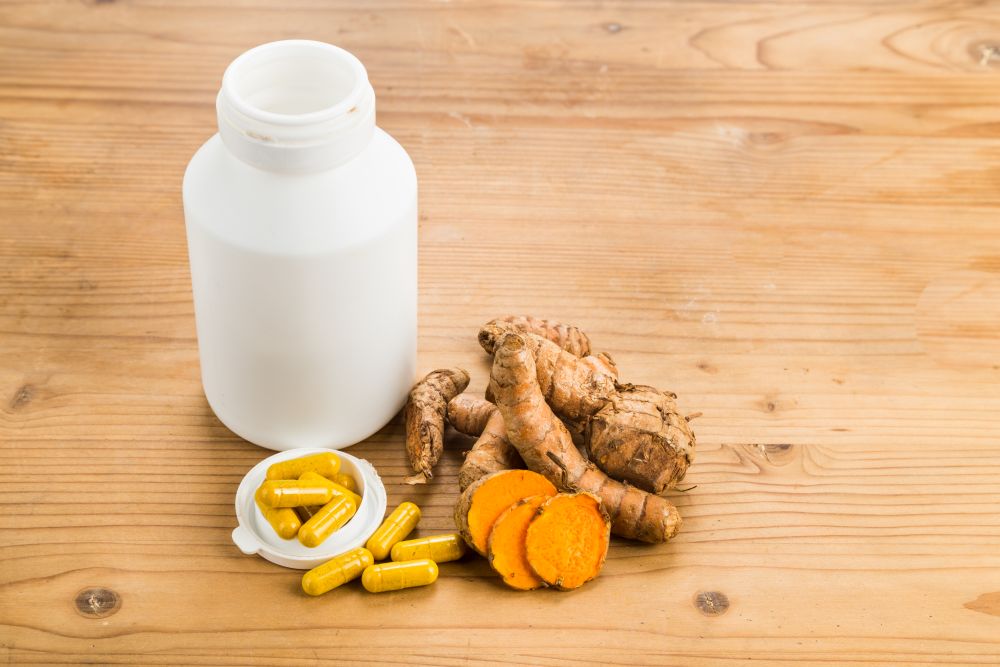 A white pill bottle, yellow capsules, turmeric roots, and turmeric slices are arranged on a wooden surface, highlighting the natural ingredients often found in a detox supplement.