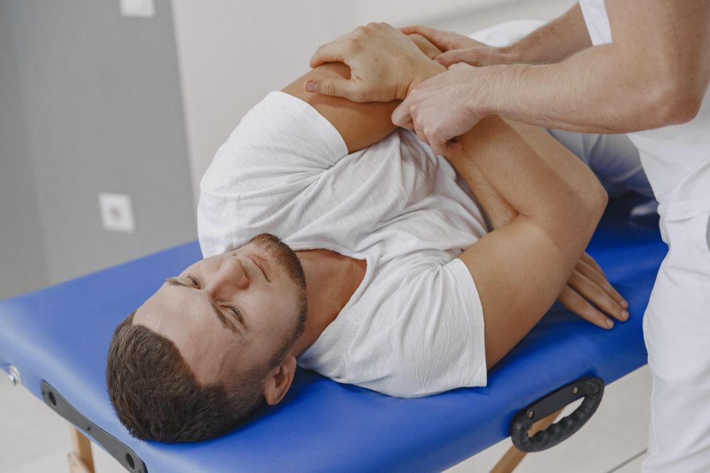 A man lies on a blue massage table while a therapist performs a chiropractic adjustment and physical therapy technique on his shoulder and arm.