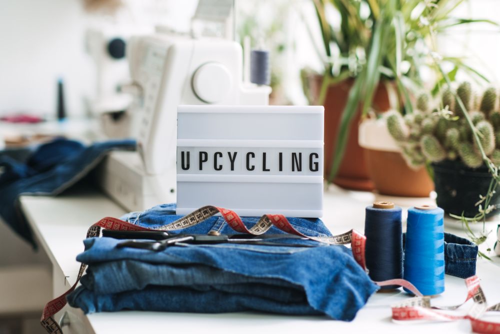 A sewing workspace with denim fabric, scissors, measuring tape, threads, and a lightbox sign that reads "UPCYCLING," surrounded by potted plants—a cozy spot for creative upcycling projects.