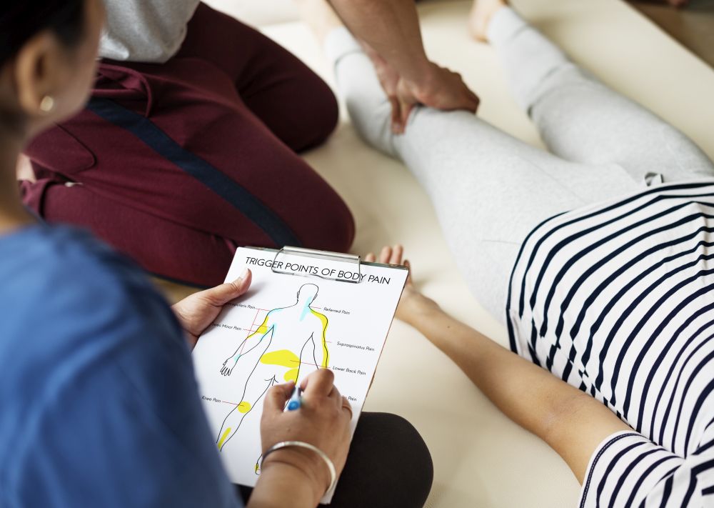 A person marks a chart titled "Trigger Points of Body Pain" while another performs a physical examination on a patient's leg at a holistic wellness clinic for inflammation and root-cause health.