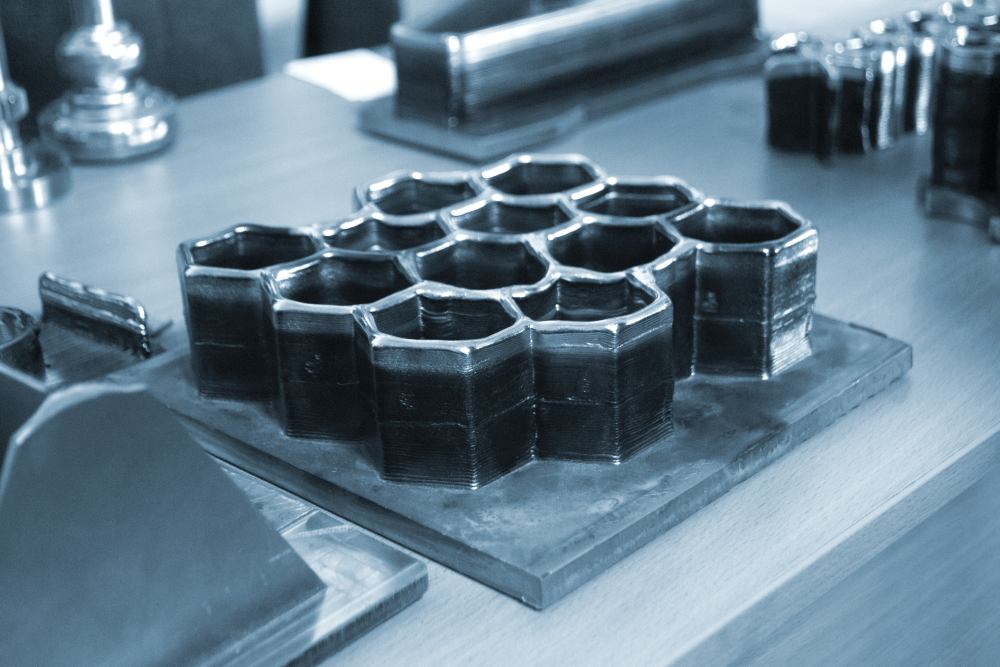 Hexagonal honeycomb-shaped black objects, created using Metal Injection Molding Solutions for Complex Precision Components, are displayed on a flat metal base atop a wooden table with various other items in the background.
